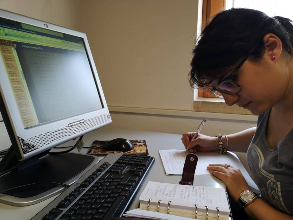 Miriam writing notes at desk with computer. Doing our Italian Ancestry Genealogy Database.